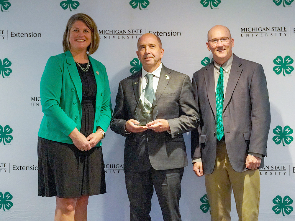 Three individuals at a backdrop. The center person holds an award.